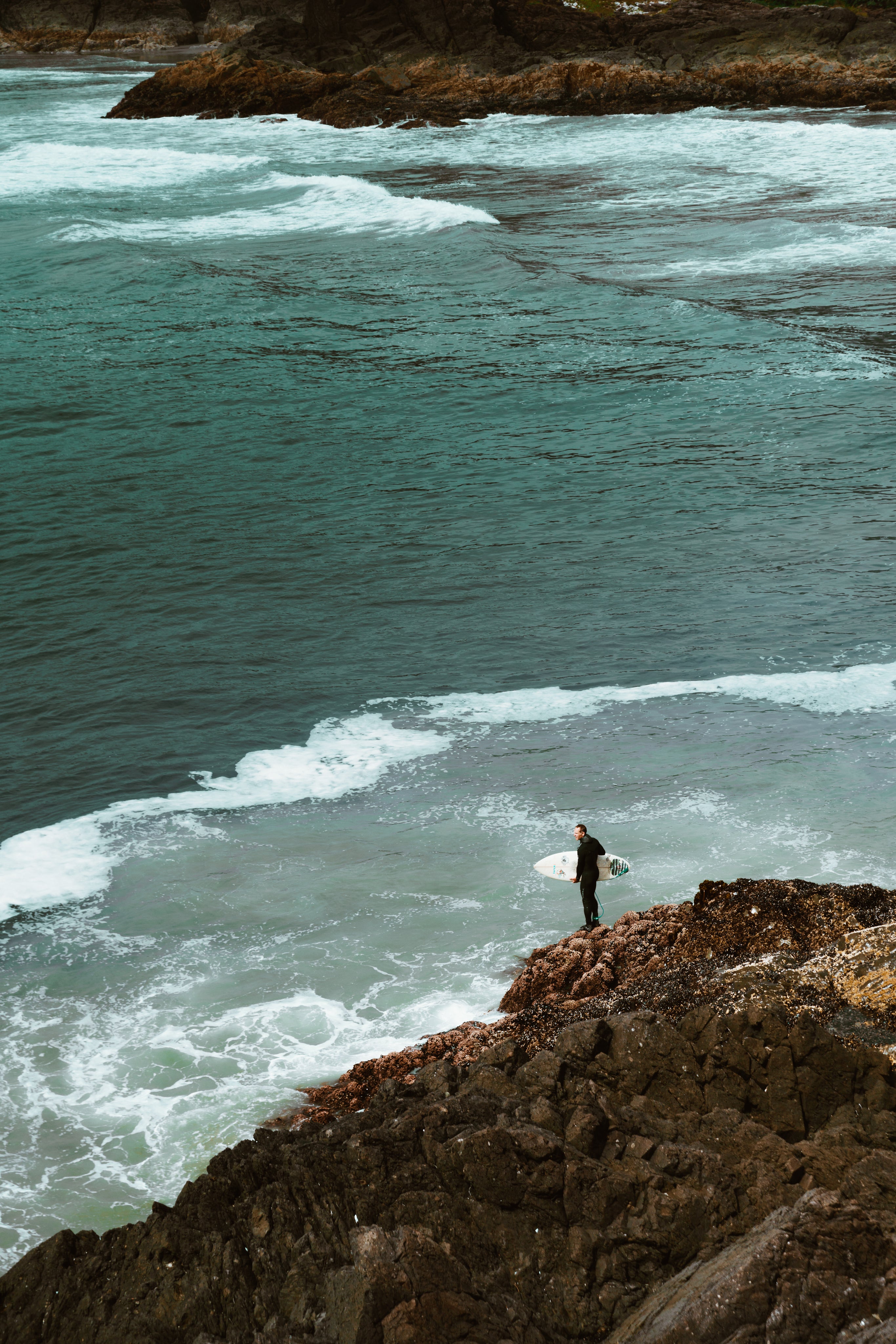 A man holding a surfboard looks out to the sea from a cliff top.